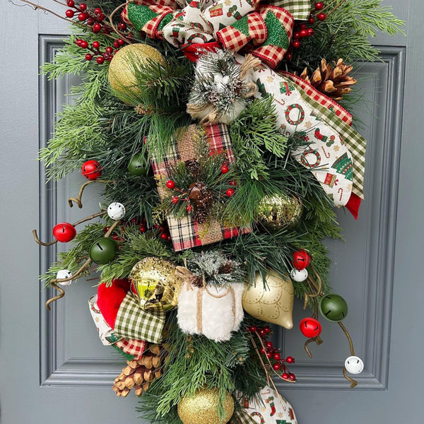 Christmas pine rustic swag with a large bow with four different ribbons. Showcases a plaid gift wrapped present with gold ornaments, pinecones, red berries, twigs and red, green and white jingle bells.